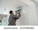 Male worker wearing uniform installing air conditioner in apartment during summer season, man technician standing indoors repairing HVAC system, checking and replacing AC filter