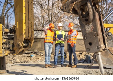 Male Workers And Construction Engineers In Hardhats Talking Near Bulldozer And Excavator. Excavator Operator. Safety Uniform Excavation Water Drainage At Construction Site.