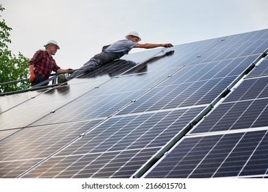 Male Workers Mounting Solar Modules For Generating Electricity Through Photovoltaic Effect. Two Men Solar Technicians In Safety Helmets Assembling Solar Panels Under Blue White Sky.