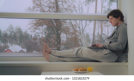 Male working with pc in flat in the morning weekend. guy wearing in casual hoodie at home typing on laptop. On the street snowing. Beautiful winter nature view with trees and pine.
