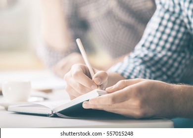 Male Writing In Notepad Placed On Desktop With Coffee Cup, Supplies, Equipment And Other Items. Close Up