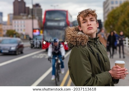 Male young adult teenager wearing a parka jacket outside drinking coffe by a red London bus, cars, a cyclist, pedestrians and traffic