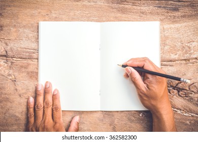 Male's Hand Holding Pencil And Writing Notebook On Old Wooden Table