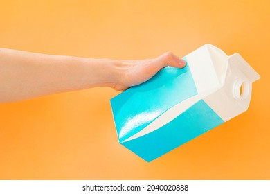 Male's Hand Tilts A Blue And White Package Of Milk To Pour A Drink Somewhere On Orange Background. Action. Open Cup