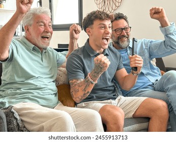 Males from multigenerational family sitting together at home looking at tv enthusiastically cheer athletes of Olympic games. Sports and participation of three attractive men of different ages - Powered by Shutterstock