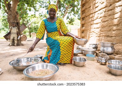 Malian housewife sourrounded by plates with food, pots and kitchen utensils cooking a meal for her family outside her house