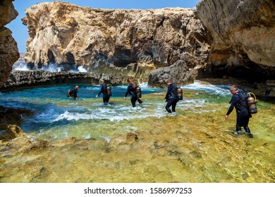 Malta Valletta June 16, 2019: A Lot Of Scuba Divers In The Equipment Are Entering The Blue Pure Water Of A Small Canyon On A Sunny Day. The Canyon Is Surrounded By Big Stones.