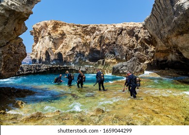 Malta Valletta June 16, 2019: A Lot Of Scuba Divers In The Equipment Are Entering The Blue Pure Water Of A Small Canyon On A Sunny Day. The Canyon Is Surrounded By Big Stones.