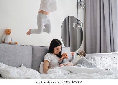 Mama with two kids at home in bedroom. Baby infant resting on mothers hands, while older sister playing, action, jumping on the bed near, Cuddle a baby while talking to toddler daughter
