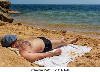 A Man 30 - 40 Years Old In A Cap, Swimming Trunks And Sunglasses Lying Sunbathing On A Sandy Beach And Looking At The Sea On A Hot Summer Day