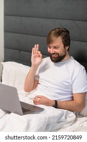 Man 30 Years Old Millennial With A Beard In A White T-shirt Sits At A Laptop Lies In Bed And Talk On Webcam Having Video Call And Shows Ok. Talks With Relatives And Friends Using Computer Webcam 