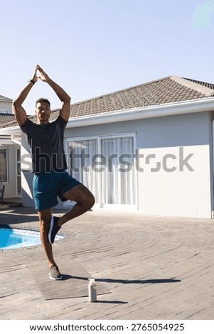 Man in 30s practicing tree pose on mat at poolside with water bottle and smartwatch. Yoga, outdoor, balance, fitness, patio, sunlit, residential
