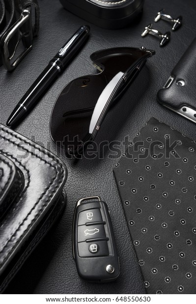 Man\
accessories in business style, sunglasses, clothes, gadgets,\
jewelry and other luxury businessman attributes on leather black\
background, fashion industry, selective\
focus
