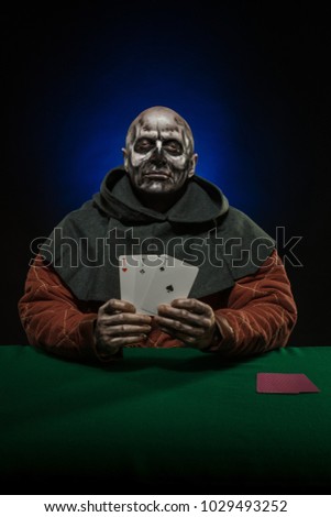 Man actor in the costume of a medieval inquisitor with a bald skull playing poker at a card table with a green cloth