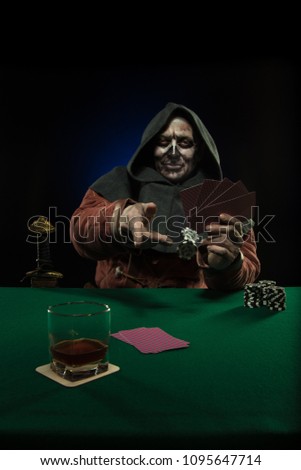 Man actor in the costume of a medieval inquisitor in a hood on his head playing poker at a card table with a green cloth