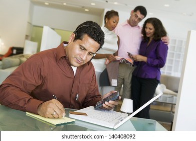 Man Adding Figures With Calculator And Laptop With Salesperson And Couple In Background At Furniture Store