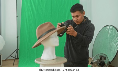 Man adjusting camera to shoot brown hat on white mannequin head against green screen in studio; creative focus.