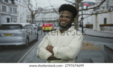Man adjusting glasses while smiling with arms crossed on a city street lined with parked cars and leafless trees; confidence friendliness.