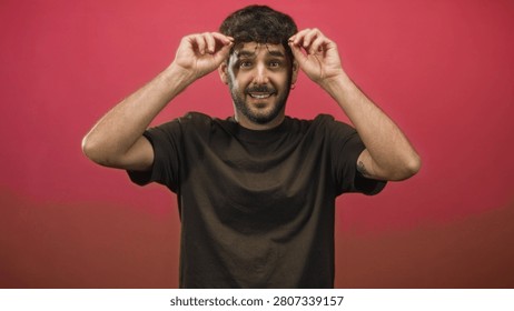 Man adjusts glasses while smiling in a studio portrait with a pink backdrop and visible tattoo on forearm; playful confidence.