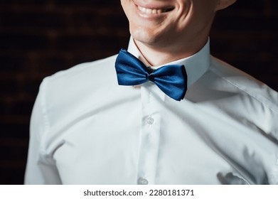 man adjusts his blue bowtie in a white shirt. attractive young man is standing in a room.