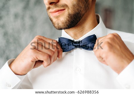 man adjusts his blue bowtie in a white shirt. attractive young man is standing in a room. closeup photo of bowtie