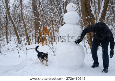 A man adjusts a large snowman while a small dog walks nearby in a snowy forest. Outdoor winter activity showing companionship and handmade snow sculpture.