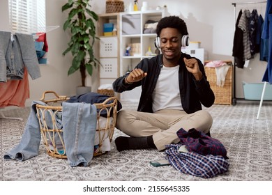 Man of African American descent is sitting on floor in middle of laundry room listening to music on wireless headphones dancing with hands folding clean clothes in background of washing machine. - Powered by Shutterstock