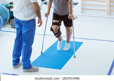 Man after car accident in an orthosis and on crutches learning to walk in the clinic, helpful therapist near him