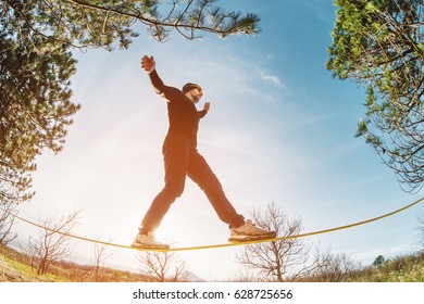 A man, aged with a beard and wearing sunglasses, balances on a slackline in the open air between two trees at sunset - Powered by Shutterstock