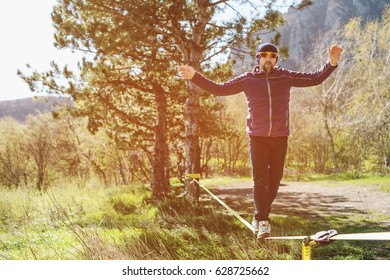 A man, aged with a beard and wearing sunglasses, balances on a slackline in the open air between two trees at sunset - Powered by Shutterstock