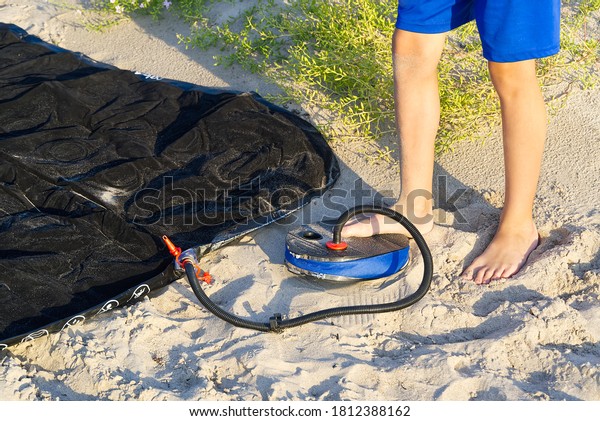 A Man with air foot pump pumps an inflatable\
mattress or air bed at sandy beach. Foot inflates air mattress with\
foot pump on sand.