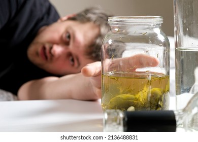 A man with an alcohol addiction lies on the bed and reaches with his hand to a jar of cucumber salted brine. Folk remedies for hangovers - Shutterstock ID 2136488831
