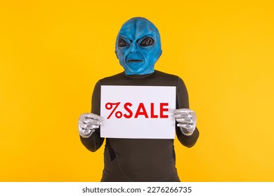 Hombre con una máscara alienígena con la cabeza azul, sosteniendo un cartel blanco con letras rojas que dice: '% de venta'. Concepto de bizarro, extraterrestre, divertido, informativo, raro y extraño.