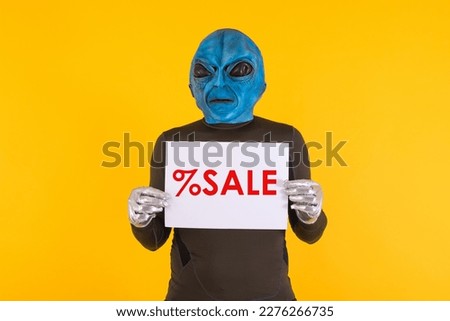 Man with an alien mask with a blue head, holding a white sign with red letters that reads: '% Sale'. Concept of bizarre, extraterrestrial, funny, informative, weird and weird.