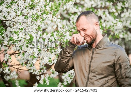 Man allergic suffering from seasonal allergy at spring in blossoming garden at springtime. Bearded young man sneezing and blowing runny nose in front of blooming tree. Spring allergy concept.