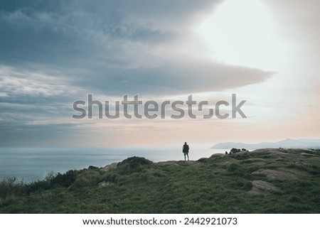 Man alone stands at the edge of a hill, looking at the sea, sun is low