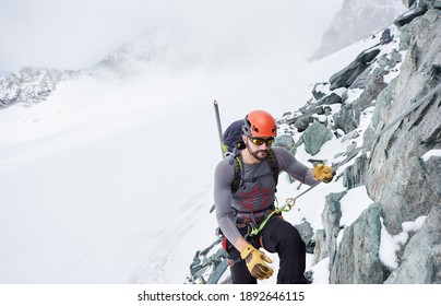 Man alpinist in sunglasses and safety helmet holding fixed rope while climbing snowy mountain. Mountaineer ascending natural rock formation. Concept of mountaineering and winter rock climbing. - Shutterstock ID 1892646115