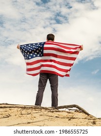 A Man With An American Flag Stands On The Sand, Skies On The Background. Man Is Holding With Both Arms Waving American USA Flag. Fourth Of July Independence Day.