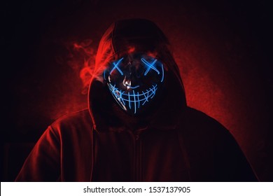 Man in angry and scary lighting neon glow mask in hood on dark red background. Halloween and horror concept.