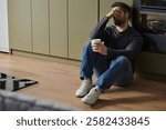 A man appears contemplative while seated on the floor, holding a cup in a stylish interior.