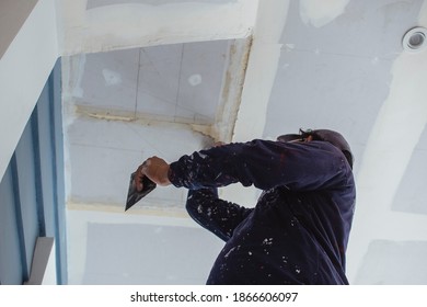 A man applies putty to a ceiling of an almost complete home. Wearing clothes full of paint splotches. Shot at the garage area.