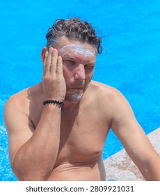 Man applies sunscreen to face while relaxing by pool. Hand with painted nails gently smears cream across skin. Bright blue water reflects sunny outdoor atmosphere
