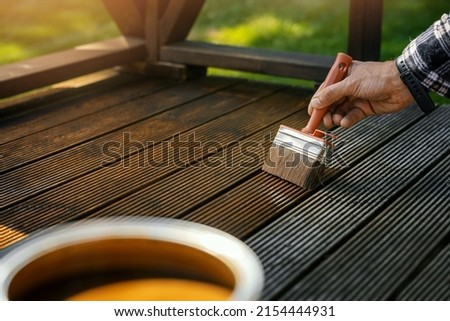 man applying brown wood protection oil on decking boards with paint brush. terrace renovation