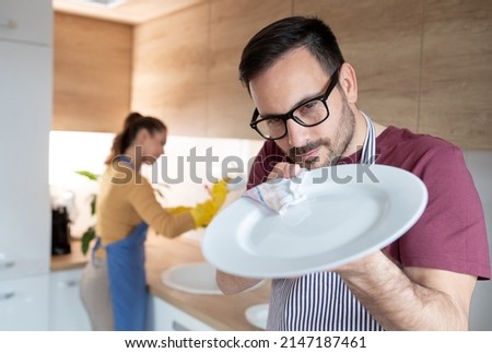 Man with apron in the kitchen polishes plate with a cloth while woman in background wash the dishes.