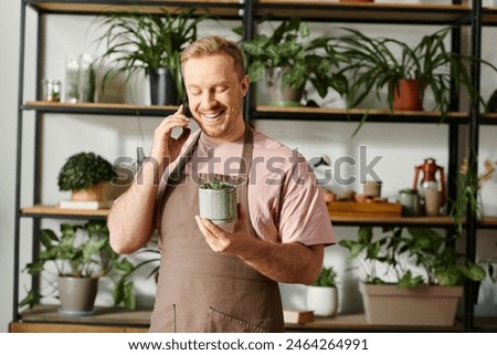 A man in an apron multitasks by holding a plant pot and talking on a cell phone in a botanical shop.