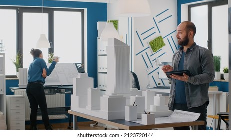 Man architect working with tablet to plan construction layout on building model. Expert contractor using maquette and digital device to design real estate structure and project.