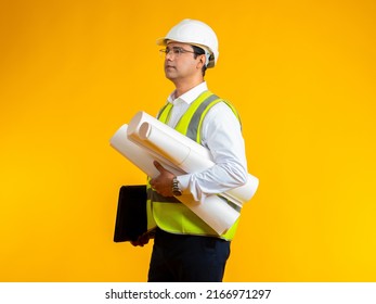 Man Architect. Young Indian Architect. Guy With Drawings In Hands On Yellow. Architect Is Hold Construction Drawings. Human In Protective Helmet And Reflective Vest. Man Employee Engineering Office