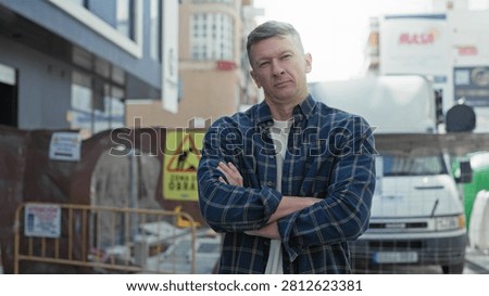 Man arms crossed in front of delivery van at a street construction site, visible hands and plaid shirt; determination.