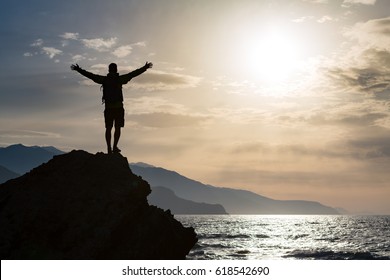 Man with arms outstretched celebrating or praying in beautiful inspiring sunrise with mountains and sea. Man hiking or climbing with hands up enjoy inspirational landscape on rocky top on Crete. - Powered by Shutterstock