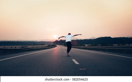 Man with arms outstretched riding a skateboard on the motorway road toward the setting sun in the background. The background is slightly blurred, focus on a skateboarder in the foreground.